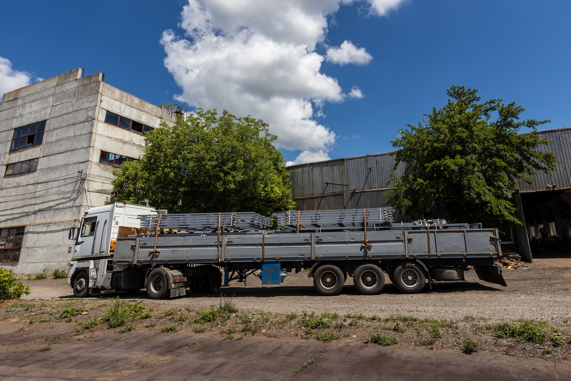 A large flatbed truck loaded with metal beams is parked near industrial buildings on a sunny day. Trees and blue sky with clouds are visible in the background. The area has a rugged, industrial vibe.